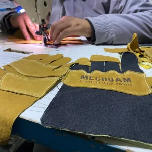 Worker assembling brown leather gloves with black reinforced cuffs at a sewing station
